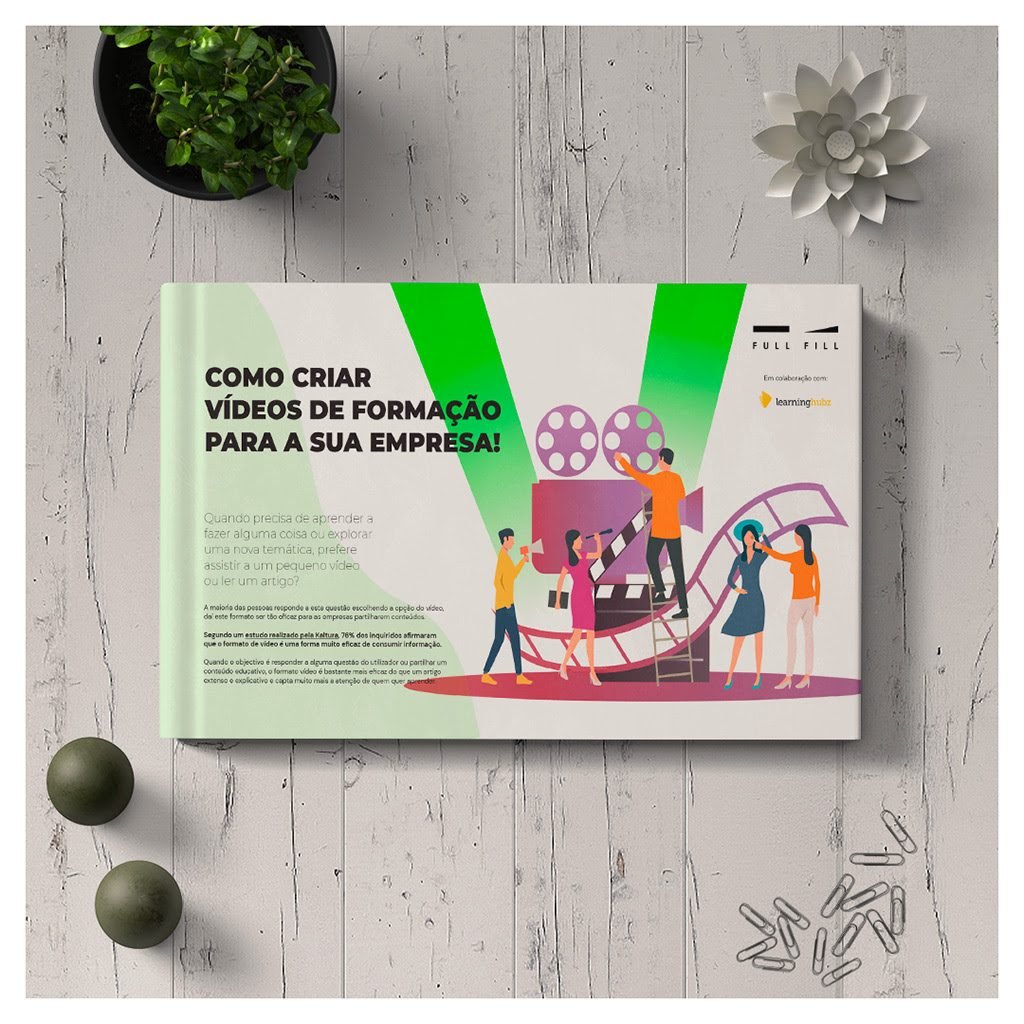 Capa do e-book «Como Criar Vídeos de Formação para a Sua Empresa», pousada sobre tábuas de madeira branca, com um vaso com uma planta, uma flor de papel e clipes espalhados à volta. A capa é clara — branco e verde-menta — com uma grande forma verde em V e uma ilustração de uma equipa a produzir vídeo.
